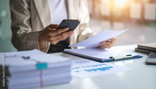A person flipping through documents on a desk and checking their smartphone