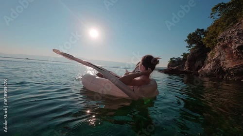 Woman smiling and enjoying golden sunset while floating on inflatable swan in calm Adriatic waters near rocky shore in Kostrena, Croatia. Summer travel and relaxation mood