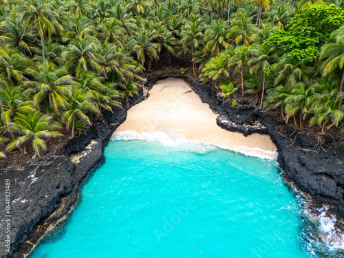 Tropical aerial view of Battery Beach on Rolas Island, São Tomé and Príncipe, showcasing turquoise water, black volcanic rocks, and dense green palm forest.
