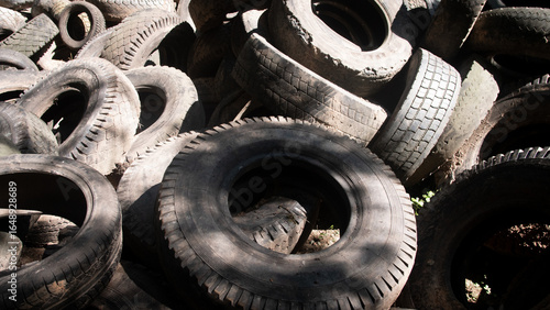 A large pile of used tires, varying in size and condition, is located in a recycling area. Sunlight filters through, casting shadows on the worn rubber surfaces.