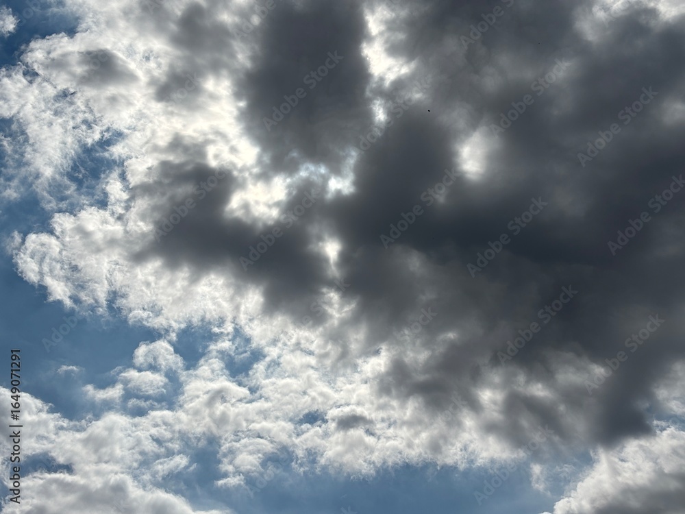 Obraz premium Bright blue sky with a smooth spread of white clouds, sunlight is seen spreading softly from behind the cloud layer in the middle, creating a calm and peaceful atmosphere.