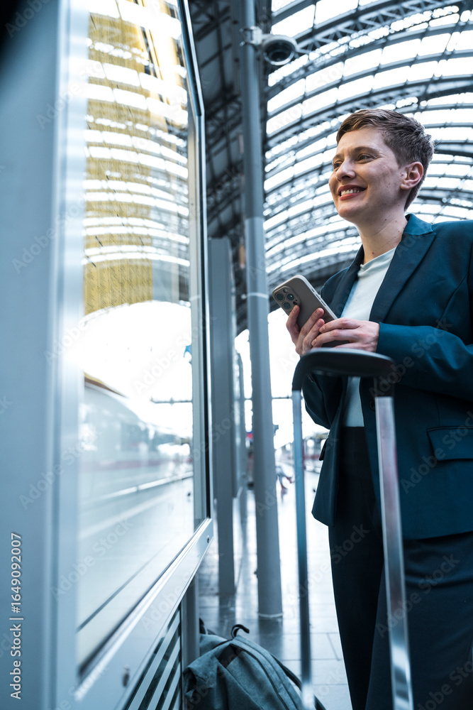 © Westend61 - Businesswoman smiling at train station with suitcase and mobile phone