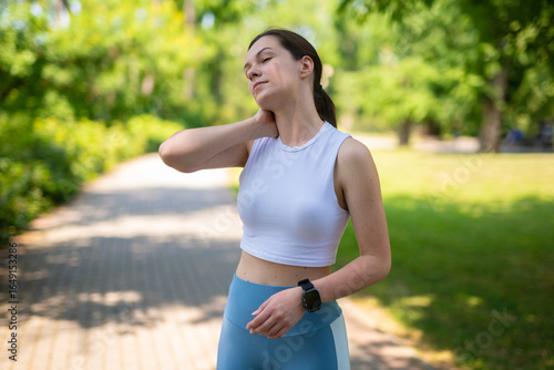 Young woman in sportswear holding her neck in pain after outdoor workout in the park.  
