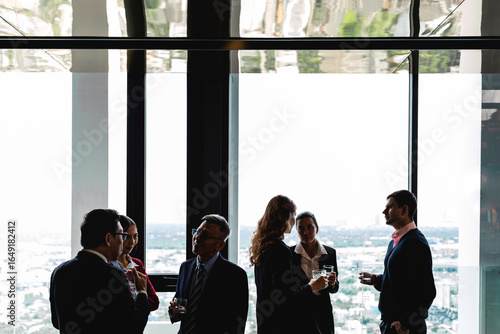 Group of diverse professionals talk casually near tall windows in a high-rise office. Relaxed after-work gathering with city views encourages conversation, networking, and team connection.