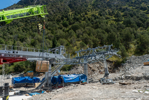 Steel Aerial cableway station frame under assembly, crane lifting crate. Pylons with sheave carriers aligned. Equipment on gravel, forest slope, construction Ala-Archa mountaineering camp, Kyrgyzstan