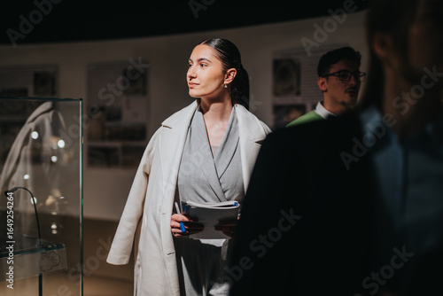 Konstfotografi Confidently dressed woman attentively observes museum exhibits, holding papers while surrounded by other visitors, creating an ambiance of curiosity and appreciation for art and history