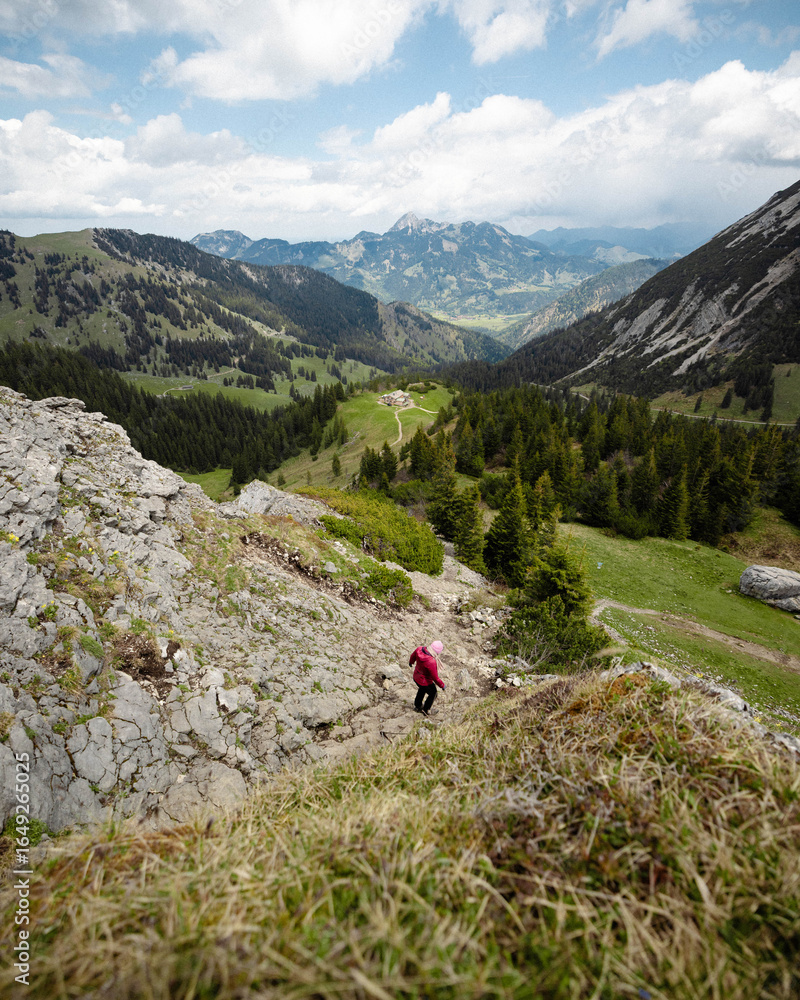 Fototapeta premium Hiker in Red Jacket Descending Rocky Mountain Slope with Scenic Valley View