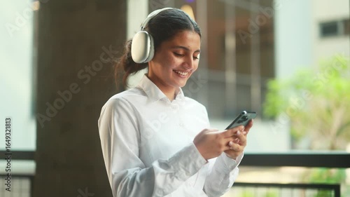 A young professional is happily smiling while enjoying her smartphone and listening to music with headphones on
