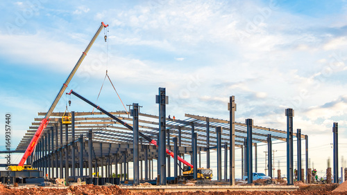 Large factory industrial building framework with workers and crane trucks are lifting metal roof beam to installing on top of warehouse structure in construction site against blue sky background