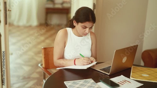 A dedicated student diligently writing notes at her desk using a laptop in a comfortable and cozy indoor setting
