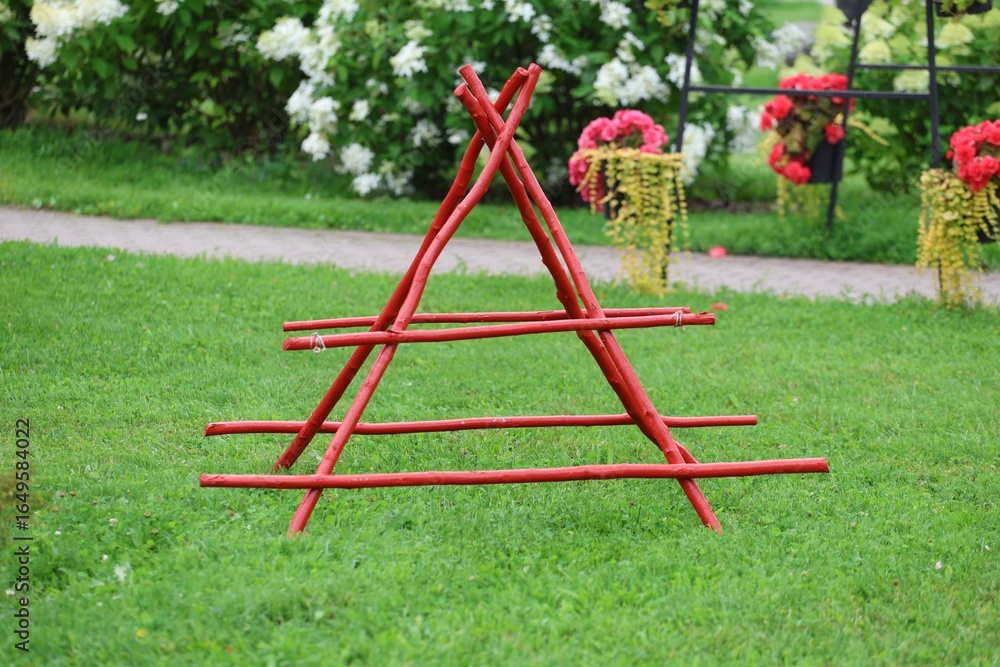 Triangular red wooden garden structure standing on lush green grass with flowers in background. Triangular red wooden garden structure standing on lush green grass with flowers in background.