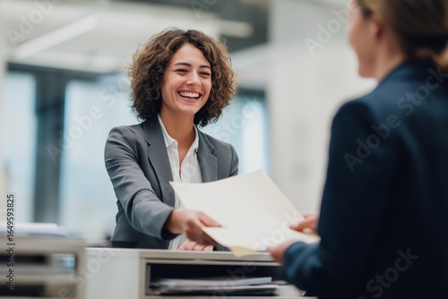 A smiling woman, wearing a gray suit jacket, is handing a file folder to a colleague in a bright office environment.