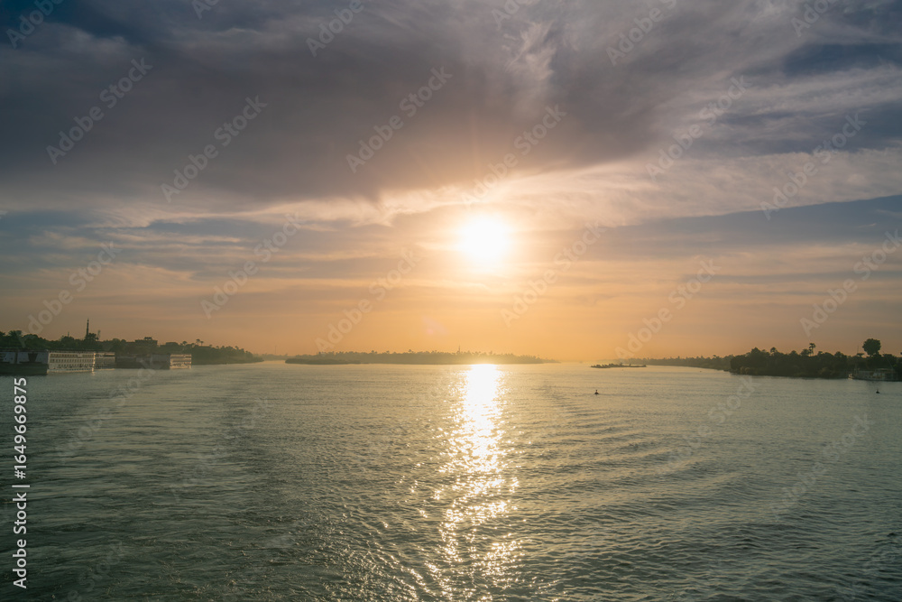 Naklejka premium Warm sunset light reflects on the Nile River near Esna Lock, Egypt, with boats, buildings, and trees lining the riverbanks.
