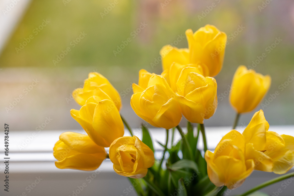 Obraz premium Close up photo of a fresh yellow tulips in a glass vase placed on a windowsill. Natural light highlights the vibrant petals and lush green leaves, creating a bright and cheerful spring mood.