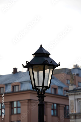 Wallpaper Mural A vintage black street lantern stands against blurred urban buildings and pale sky, capturing the elegance of classic design within a modern city landscape. Torontodigital.ca
