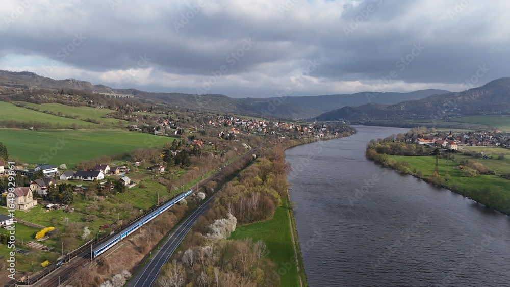 Fototapeta premium Drone view of a winding river with a passing train, green hills, and small villages. Scenic countryside landscape with mountains in the background under dramatic cloudy sky.