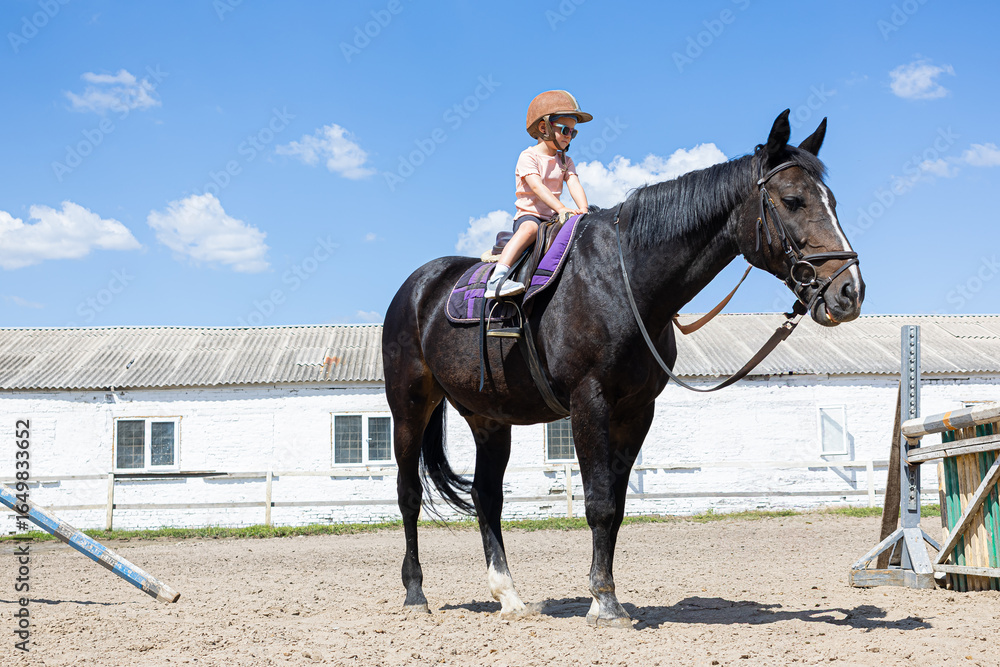 Smiling little girl riding a black horse at equestrian training ground Smiling little girl riding a black horse at equestrian training ground