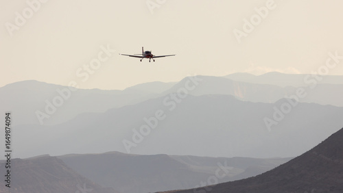 A small airplane flies towards the camera with layers of distant mountains hazy with dusty morning light in the background.