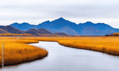 A winding river snakes through a field of golden grasses toward imposing, blue-hued mountains beneath a cloudy sky. Autumn colors dominate the landscape