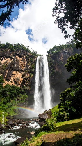 Tall waterfall cascading down rocky cliffs, a rainbow at its base, lush green vegetation surrounding it