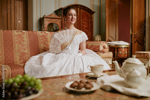 Caucasian young adult woman sitting on ornate sofa holding lace fan wearing historical dress, smiling slightly with hair styled up table with tea set and fruit in foreground