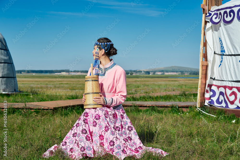 Fototapeta premium A Mongolian girl wearing Mongolian attire and holding a bucket of milk tea in front of a yurt.