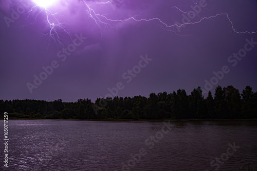 Bright purple lightning streaks across a stormy night sky above a dark forest and calm lake.