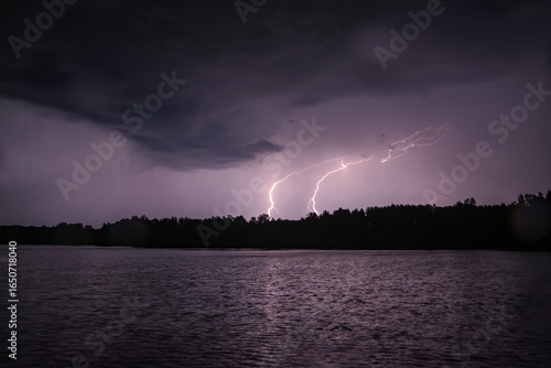 Fototapeta Naklejka Na Ścianę i Meble -  Lightning storm over a dark lake with forest silhouette under dramatic night clouds.