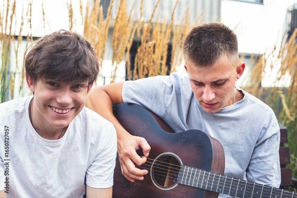 Fototapeta premium Two young people relaxing outdoors with a guitar. A young man is playing the guitar while a teenage boy listens cheerfully. A warm and joyful moment of friendship and music.