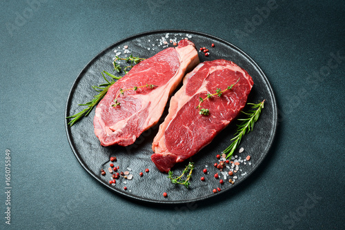 Two raw rib eye beef steaks with spices on a plate. Ready to cook. On a dark stone background. Top view.