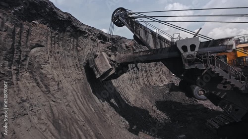 Concept mining industry from FPV drone view. Walking excavator removes overburden soil and loads it into large yellow dump truck at coal quarry.