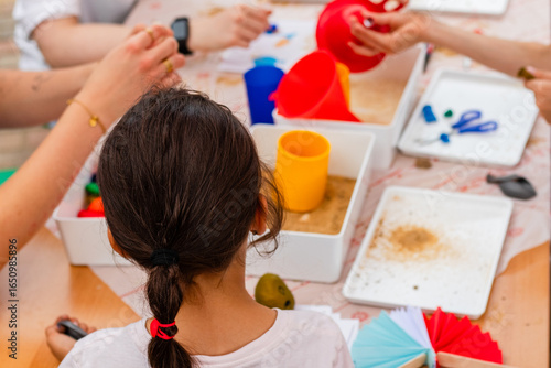 Young girl with dark hair, engaged in creative activities at an after-school program, surrounded by colorful materials and hands of peers, fostering collaboration and artistic expression