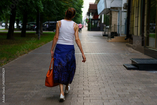 Woman walks down a city sidewalk in a blue skirt and white top during a sunny afternoon in the urban setting