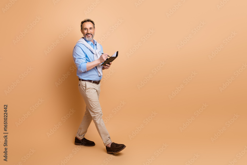 Mature businessman walking confidently holding a notebook in stylish attire against a beige background Mature businessman walking confidently holding a notebook in stylish attire against a beige background