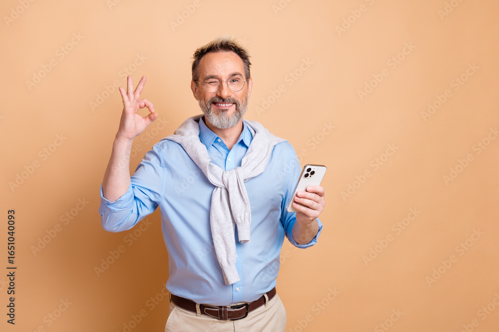 Confident mature businessman with glasses gesturing OK sign and holding a smartphone against beige background Confident mature businessman with glasses gesturing OK sign and holding a smartphone against beige background