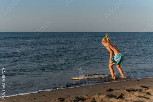 Shirtless young blond boy with hair in a bun playing at the shoreline, wearing blue floral shorts, leaning forward toward skimboard as it splashes in shallow ocean water on sandy beach