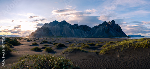 Fototapeta Naklejka Na Ścianę i Meble -  The Vestrahorn mountains rise against a dramatic sky, while green grass peeks through black sand dunes, highlighting Icelands breathtaking beauty at sunset.