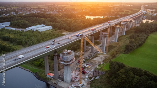 Highway bridge construction site over the Kiel Canal, with traffic moving on the existing lanes while large cranes and construction equipment work on building the new bridge pillars and the roadway.