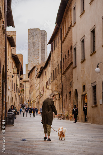 Woman in olive coat walking her Cavalier King Charles Spaniel along a picturesque medieval cobblestone street in San Gimignano, Tuscany, seen from behind