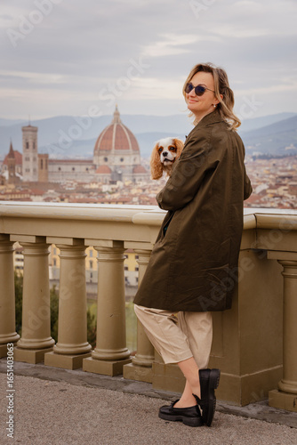 Schilderij op canvas Stylish woman holding a Cavalier King Charles Spaniel on a scenic terrace overlo