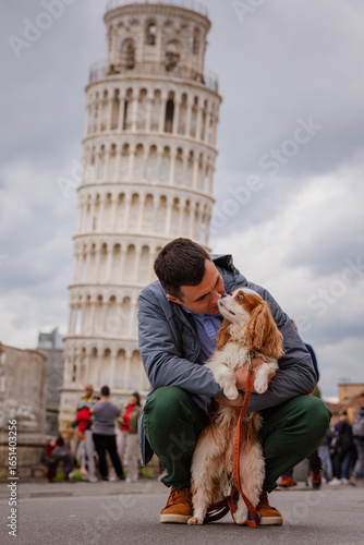 Young man crouching and bonding with a Cavalier King Charles Spaniel dog in front of the famous Leaning Tower of Pisa, Italy