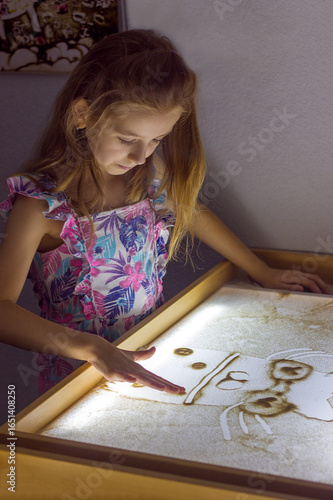 Beautiful little girl draws with sand on a light square, art therapy, sand animation, creative leisure for children, selective focus