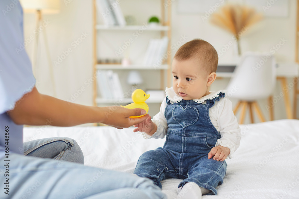Mother and baby playing with duck toy, sitting in bed at home. Mom learning child through interaction and game, hand of woman giving duck to curious infant kid. First skills, development concept Mother and baby playing with duck toy, sitting in bed at home. Mom learning child through interaction and game, hand of woman giving duck to curious infant kid. First skills, development concept