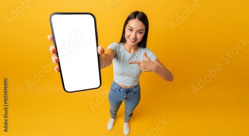High angle view of a joyful young Japanese woman presenting a blank smartphone screen for your app mockup on a vibrant yellow studio background.