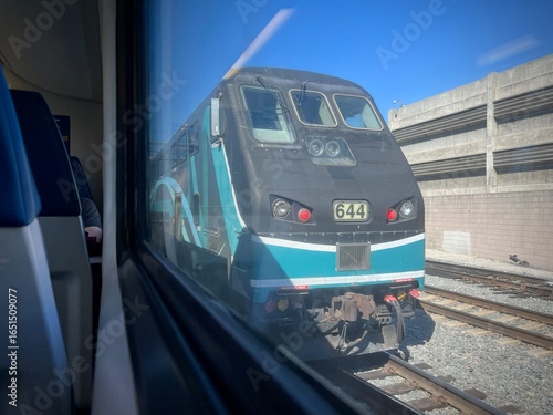 LOS ANGELES, CA, APR 19, 2025: view from inside a train showing Metrolink locomotive in teal and gray colors on parallel tracks, with concrete infrastructure and clear blue sky visible