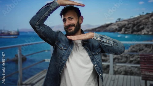 Hispanic man with beard, in denim shirt, making a gesture by the beach with the sea and mountains in the background, capturing a relaxed and joyful outdoor moment.