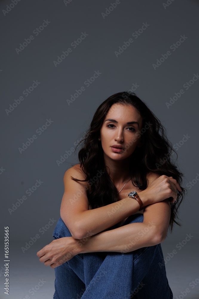 Naklejka premium Beautiful tall thin brunette with curly hair in the studio, cyclorama. The girl stands shirtless covering her nakedness with her hands, gray background. Studio photo shoot for a model. Portrait. 