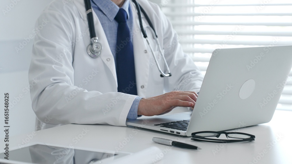 Male doctor in lab coat and stethoscope sitting at desk typing on laptop, holding glasses and pen nearby in medical office. Medicine and healthcare concept