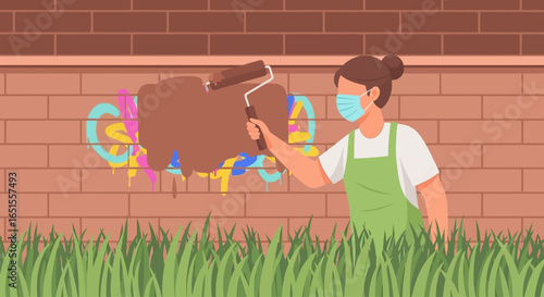 A person wearing a face mask and apron paints over colorful graffiti on a brick wall with a roller.