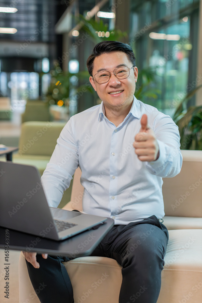 Naklejka premium Like. Happy young businessman guy working on laptop looking approvingly at camera showing thumbs up, like sign positive something good, positive feedback sitting in modern office lobby. Businesspeople